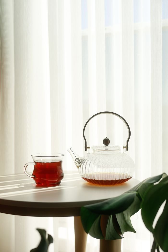 A glass teapot and teacup filled with tea on a table by sheer curtains, creating a serene morning atmosphere.