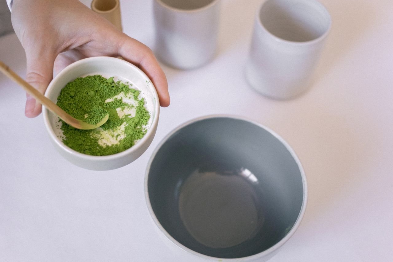 Home A hand holding a bowl of vibrant green matcha powder with a wooden spoon, ready for tea preparation.